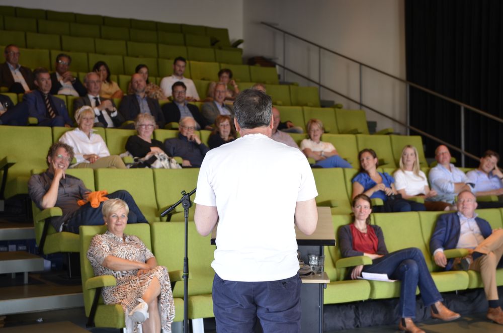 Een persoon spreekt voor een publiek in een collegezaal of auditorium. Het publiek zit op groene stoelen in rijen, en de spreker staat met de rug naar de camera. Er is een microfoonstandaard bij de spreker. De ruimte heeft een modern ontwerp met een trap zichtbaar aan de rechterkant.
