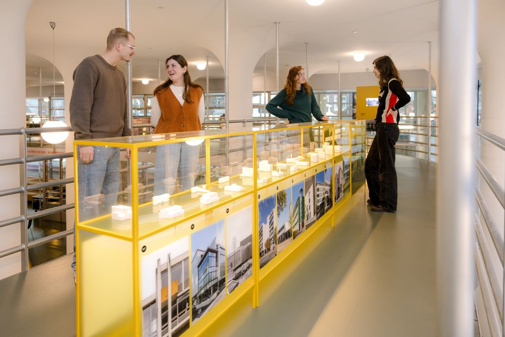Vier bezoekers staan langs een lange gele vitrine met architectuurmodellen en foto’s, in een lichte museumruimte met ronde kolommen.