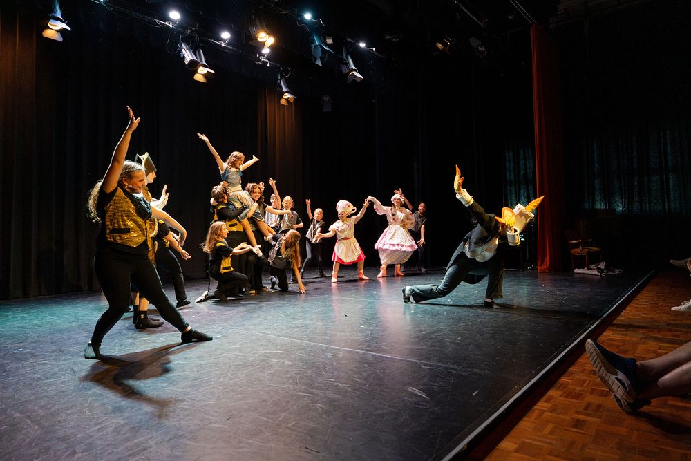 Groep performers staat op een theaterpodium tijdens een musicalscène van Belle en het Beest, met uitbundige houdingen en kostuums waaronder jurken en een hoofd met hoorns of masker. Op de voorgrond knielt een performer met uitgespreide armen terwijl de rest in verschillende posities over het podium verdeeld staat onder theaterverlichting.
