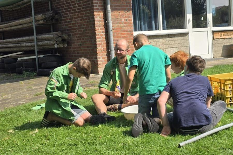 Groep kinderen zit samen met een volwassene op het gras naast een gebouw en doet een buitenactiviteit. Rechts staat een gele krat met materialen.
