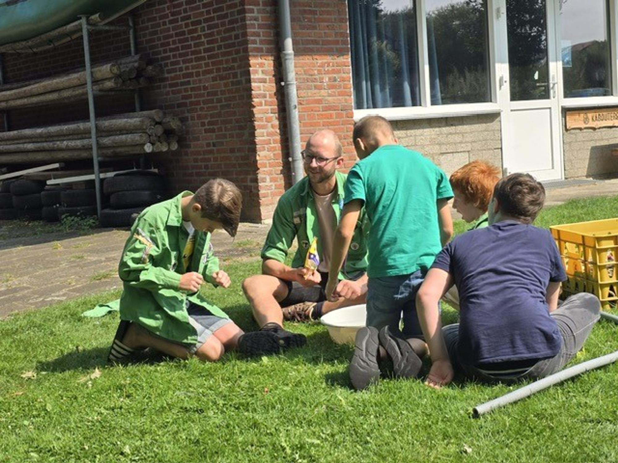 Groep kinderen zit samen met een volwassene op het gras naast een gebouw en doet een buitenactiviteit. Rechts staat een gele krat met materialen.