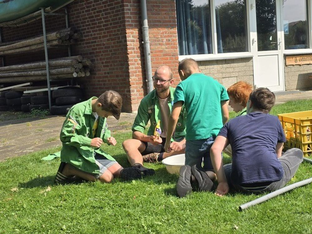 Groep kinderen zit samen met een volwassene op het gras naast een gebouw en doet een buitenactiviteit. Rechts staat een gele krat met materialen.
