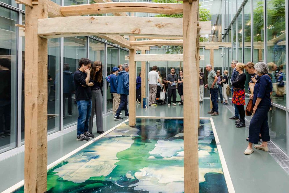 Bezoekers lopen door een installatie met houten constructies en een groot werk op de vloer in de glazen binnenruimte van SCHUNCK.