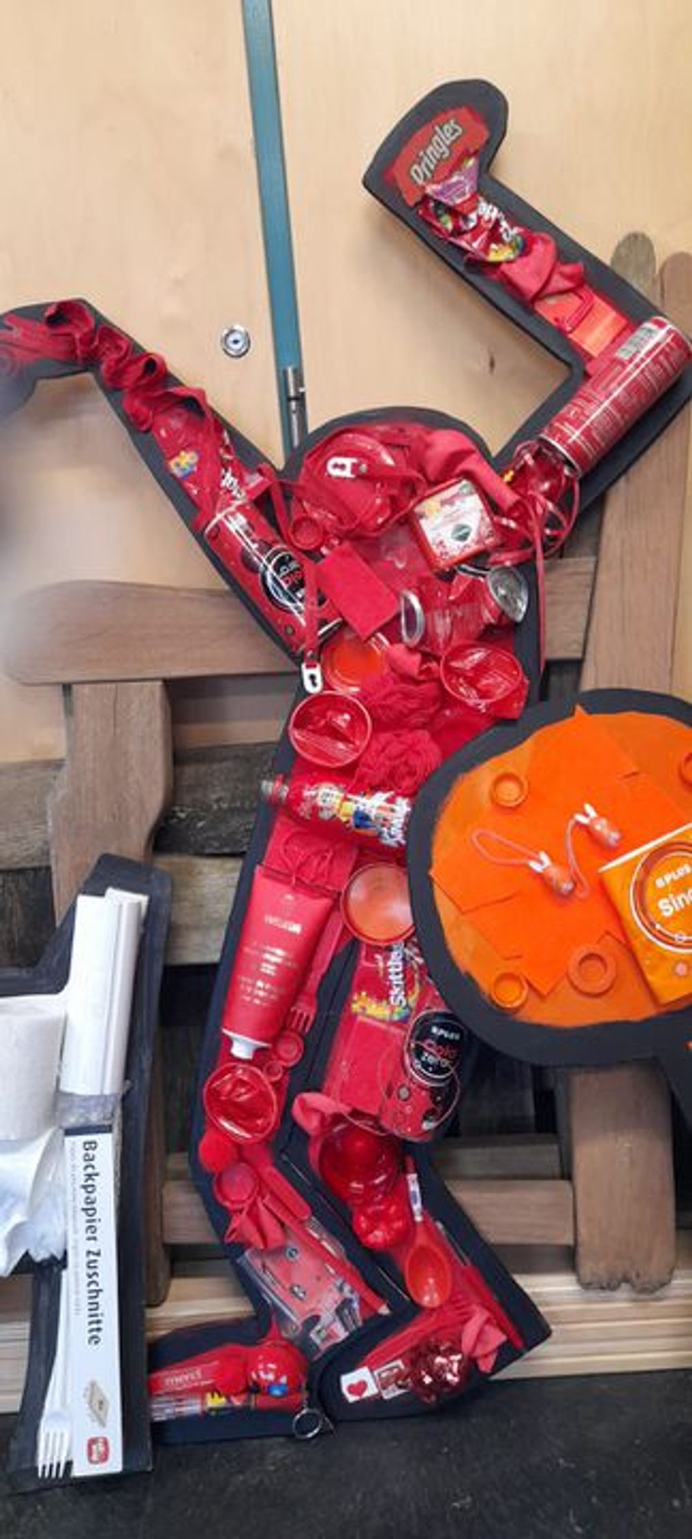 Mensfiguur gemaakt van rode objecten: De figuur is samengesteld uit flessen, doppen en andere plastic items, gemonteerd op een houten achtergrond. 