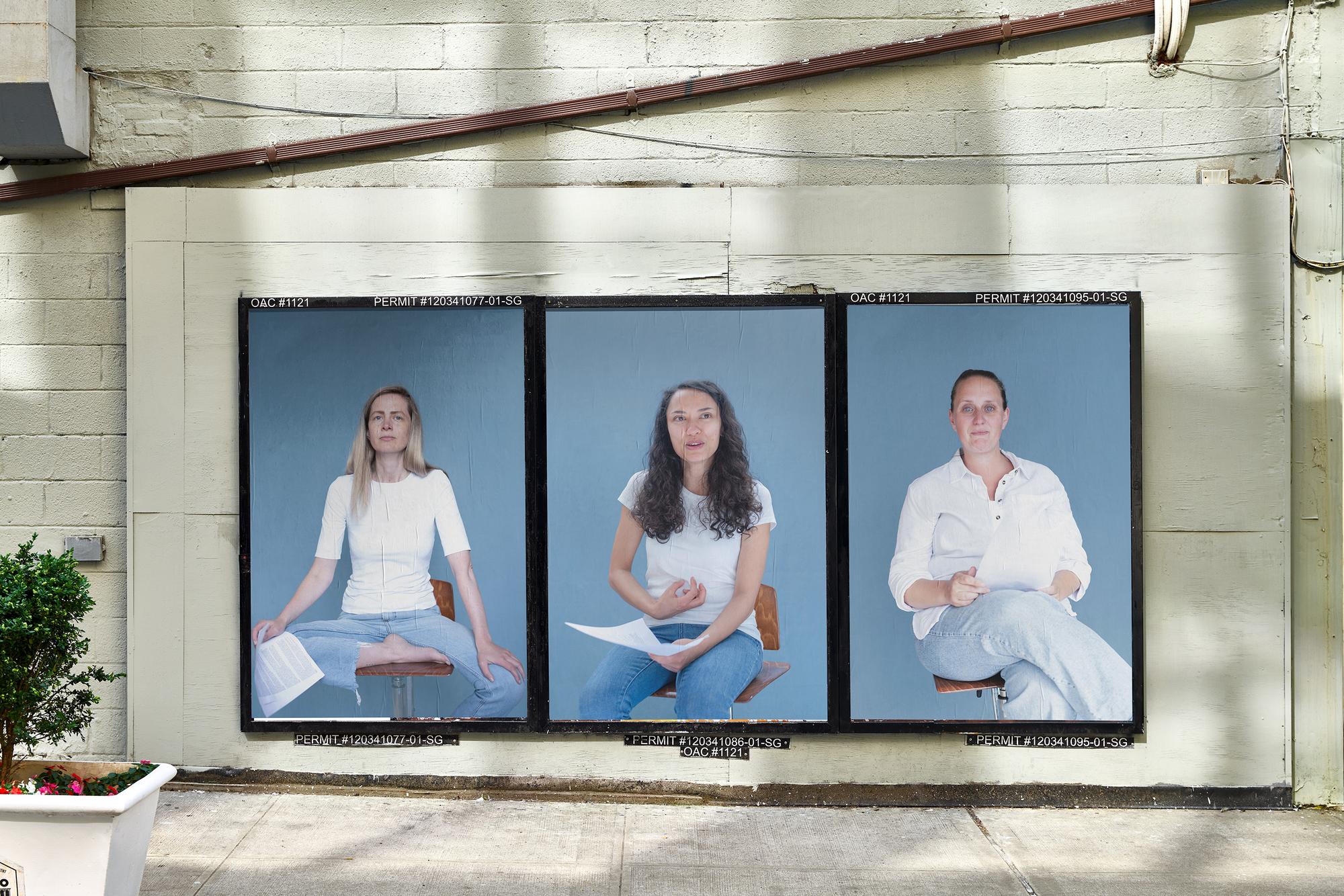 Drie grote portretfoto’s van vrouwen tegen een blauwe achtergrond hangen naast elkaar aan een buitenmuur in de openbare ruimte.