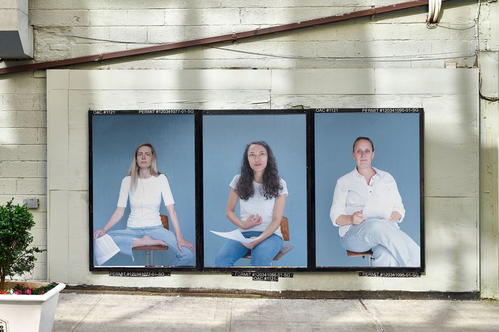 Drie grote portretfoto’s van vrouwen tegen een blauwe achtergrond hangen naast elkaar aan een buitenmuur in de openbare ruimte.