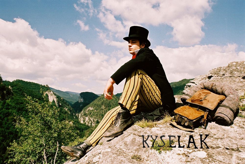 Filmstill uit Kyselak war da! waarin Joseph Kyselak in historische kleding op een rots zit met uitzicht over een berglandschap.