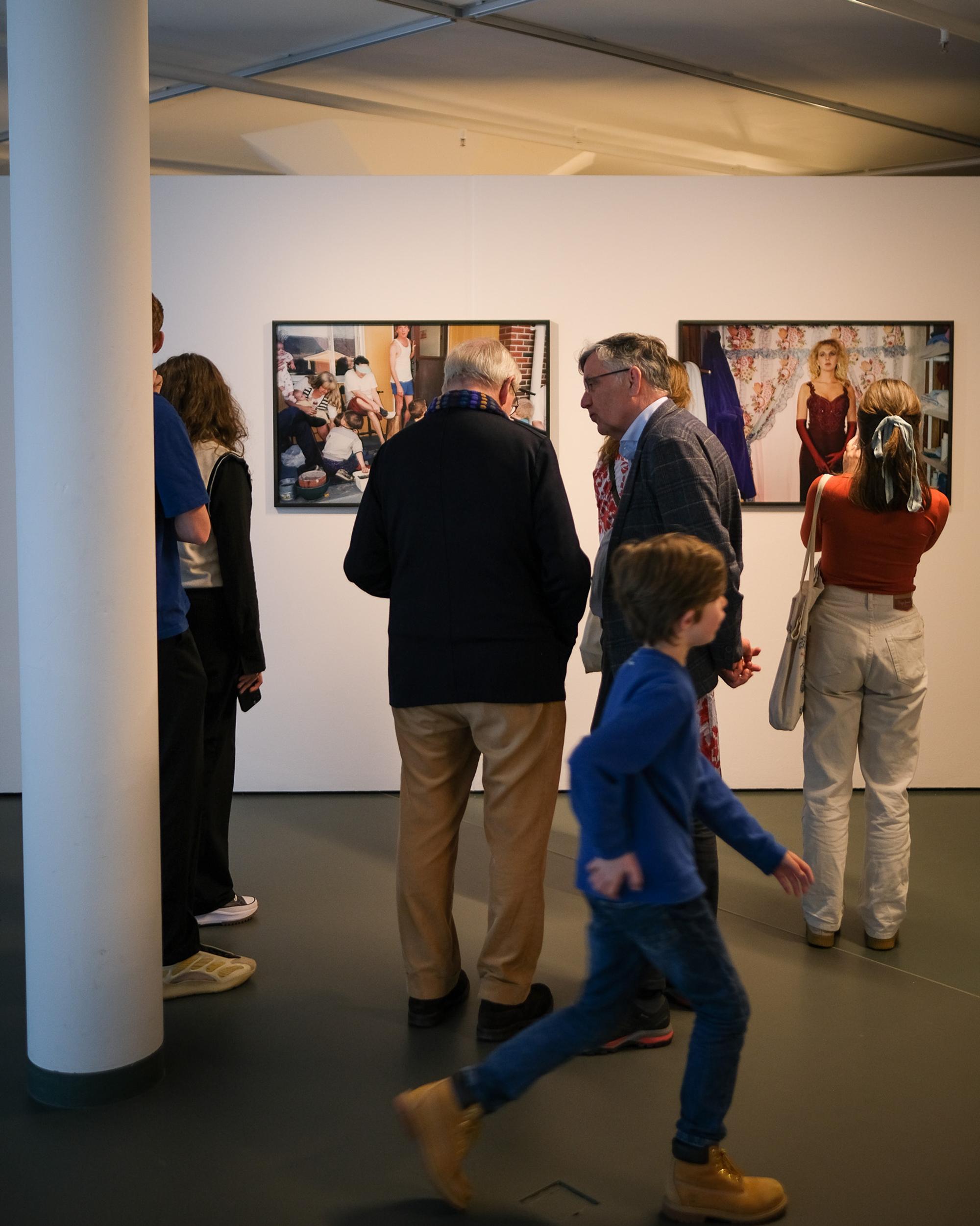 Bezoekers van verschillende leeftijden bekijken foto’s aan de muur tijdens een tentoonstelling in SCHUNCK; een kind loopt speels door het beeld.