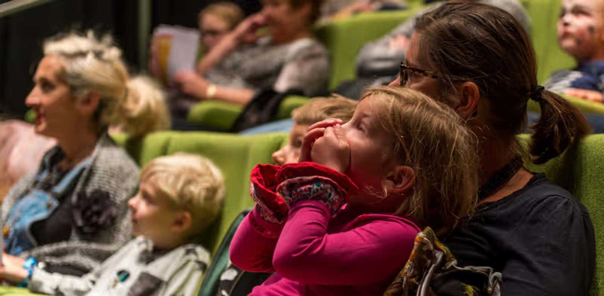 Kinderen en volwassenen zitten samen in een theaterzaal met groene stoelen en kijken aandachtig naar een voorstelling. Op de voorgrond zit een meisje in een roze trui geboeid naar het podium te kijken, omringd door andere bezoekers.
