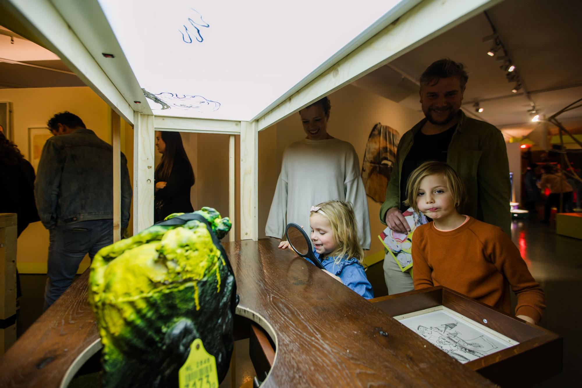 Twee kinderen kijken naar een bureau waar een nep-hoofd met gele verf doorheen komt. Een meisje heeft een spiegeltje uit de bureaula gepakt. 