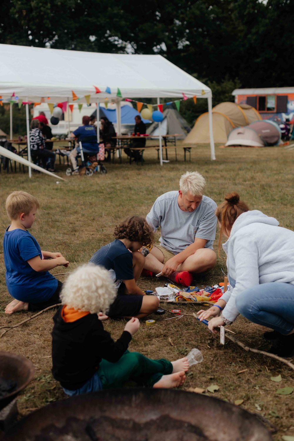 Kinderen en volwassenen knutselen samen op een grasveld tijdens een zomerkamp, met kleurrijke vlaggetjes, tenten en een partytent op de achtergrond.
