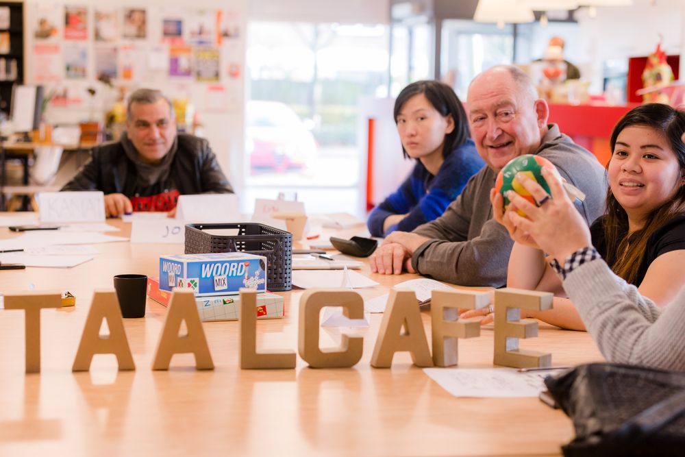 Vijf bezoekers zitten aan een tafel in het taalcafé, waar de letters 'TAALCAFE' en verschillende taalspelletjes op de tafel staan.