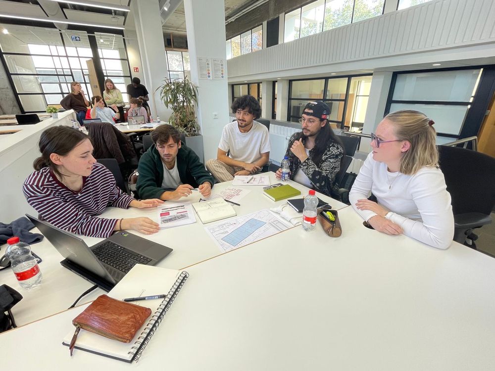 Studenten werken in kleine groepen aan architectuurplannen in een lichte ruimte. Op de voorgrond bespreken vijf deelnemers tekeningen en documenten aan een witte tafel.