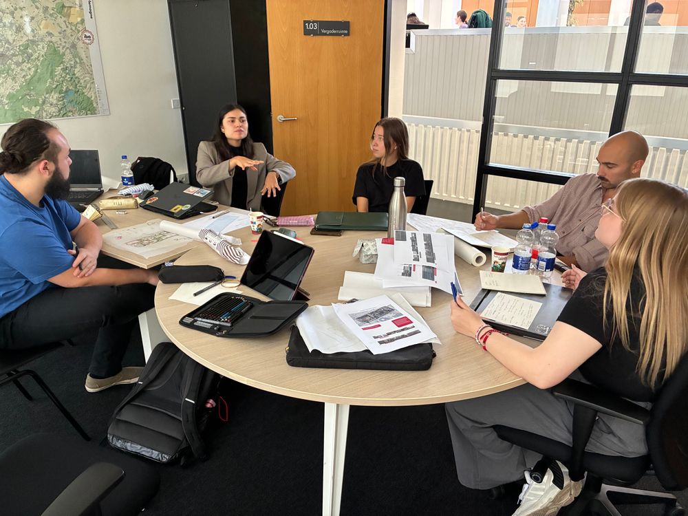 Vijf deelnemers overleggen rond een ronde tafel vol architectuurtekeningen, schetsen en laptops. Een van hen maakt handgebaren tijdens het gesprek.