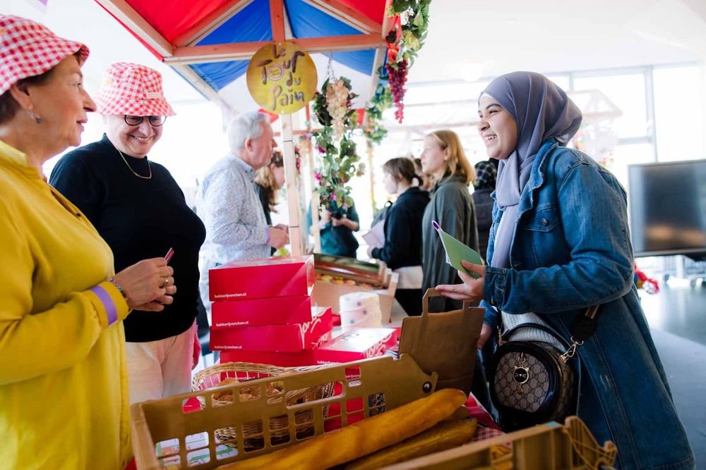Twee stands verbeelden ondernemers. Op de voorgrond een bakker waarin twee medewerksters te zien zijn die een fictieve klant ontvangen, een leerlinge die deelneemt aan Taaldorp. In de stand daarachter zijn nog twee medewerkers te zien die drie jongeren te woord staan.