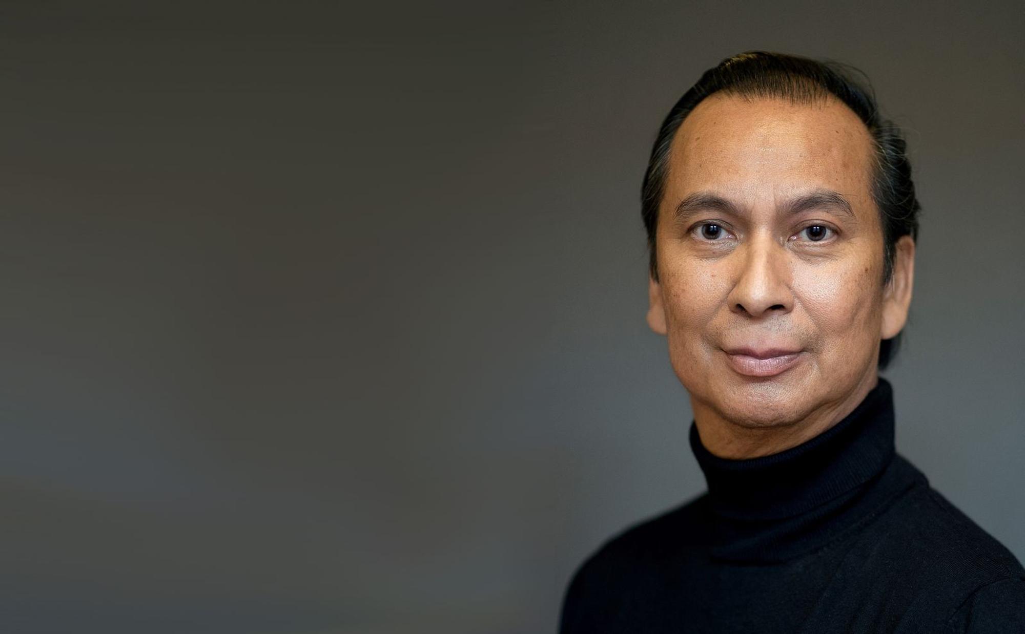 Een portretfoto van schrijver Reggie Baay die een zwarte coltrui draagt en staat voor een grijze achtergrond.