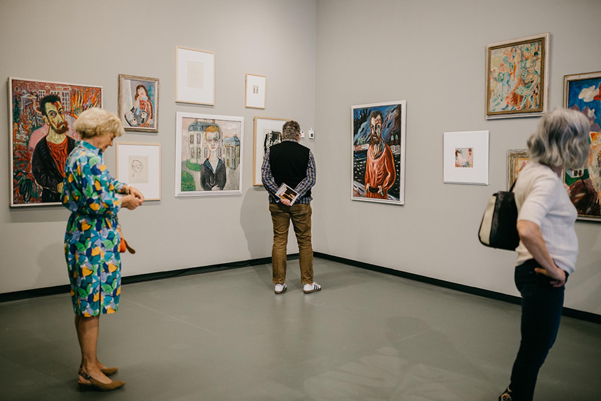Bezoekers bekijken kleurrijke schilderijen aan de muur tijdens een tentoonstelling in een museumzaal. Eén persoon staat met de handen op de rug geconcentreerd naar een werk te kijken, terwijl anderen rondlopen.