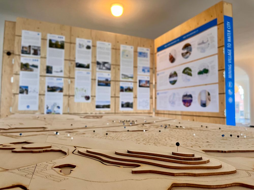 Houten maquette met markeringen, voor een houten wand met informatieve posters op de achtergrond.