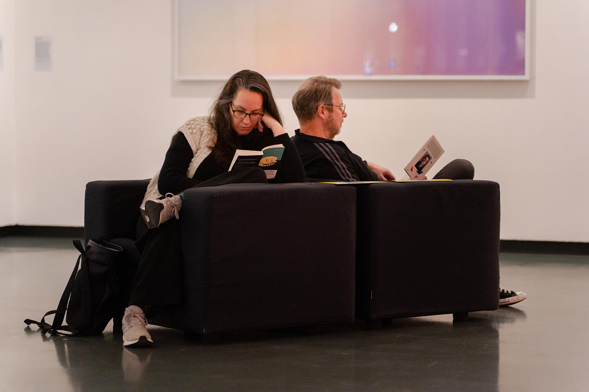 Twee personen zitten rug aan rug op lage zitbanken in een rustige binnenruimte en lezen ieder een boek. Op de achtergrond hangt een groot, abstract kunstwerk aan de muur.