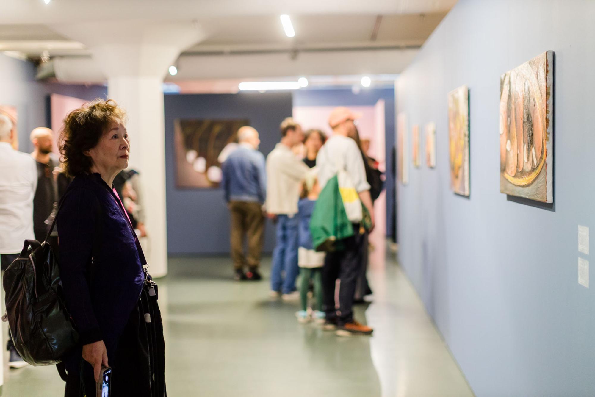 Persoon bekijkt schilderijen aan een blauwe wand in een museumruimte, terwijl andere bezoekers verderop staan.