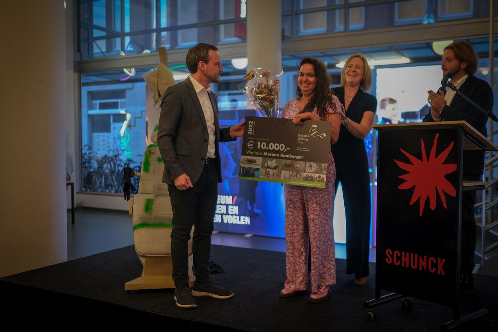 Wethouder overhandigt prijs aan Morena Bamberger op het podium in de centrale hal van het SCHUNCK Glaspaleis.