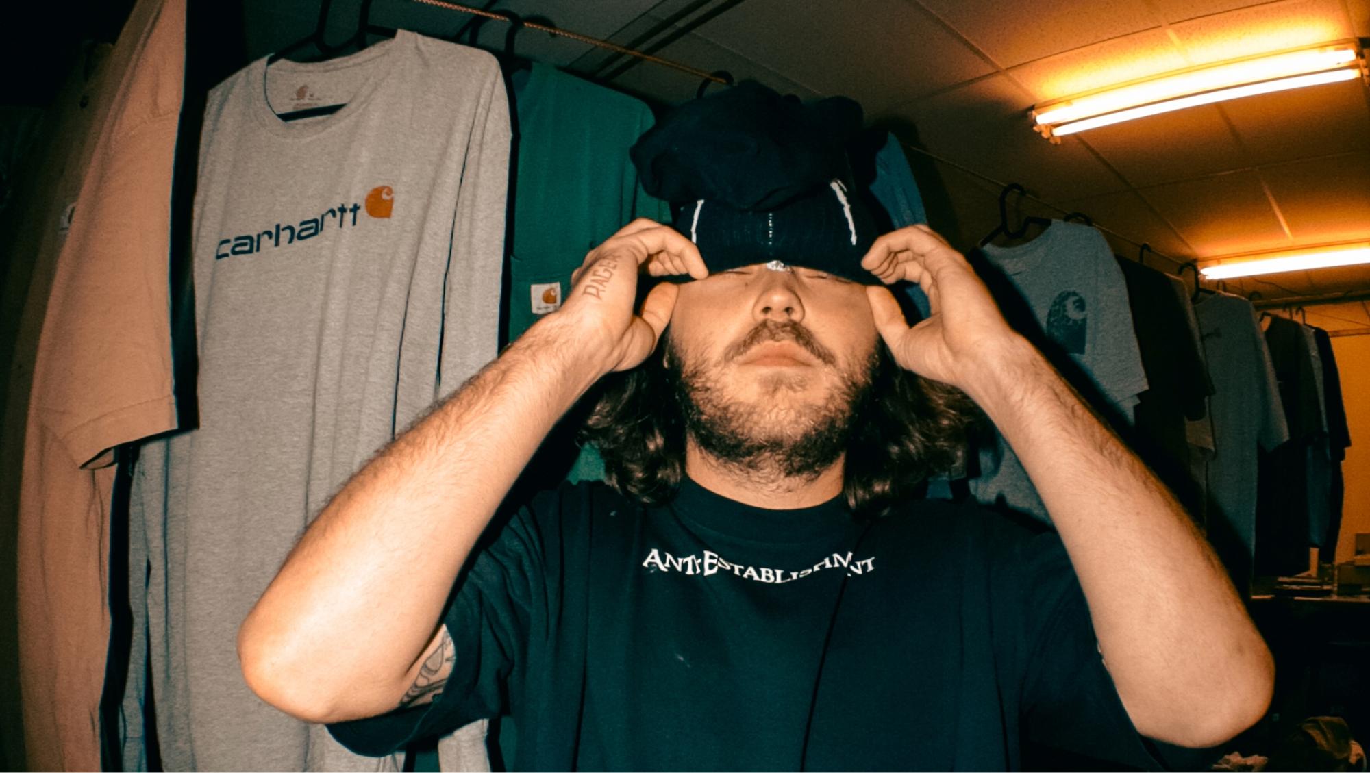 Portret Naud Verboeket van Tears. Hij trekt een muts over zijn ogen en staat voor een rek met t-shirts. 