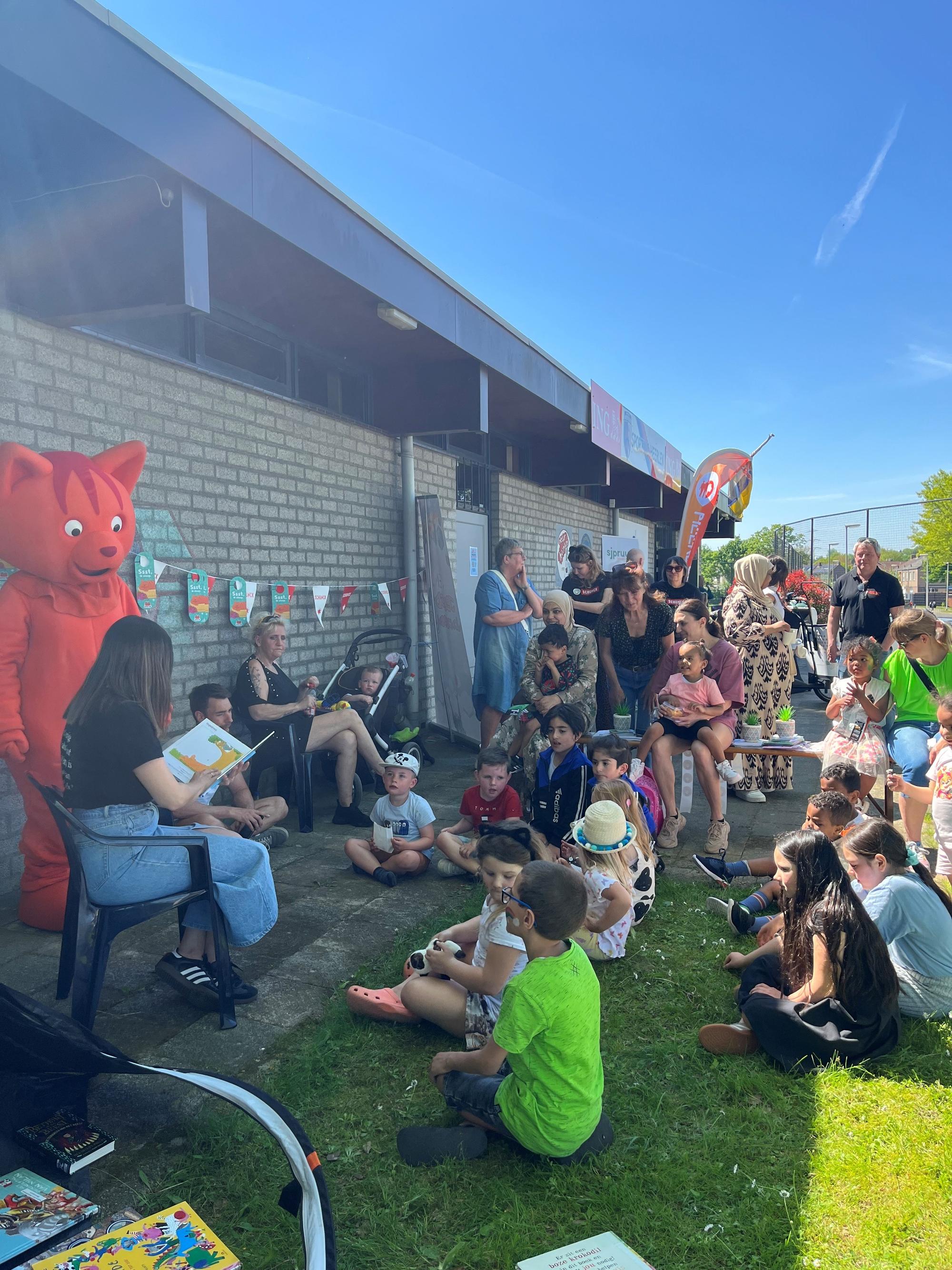 Een groep jonge kinderen zit buiten op het gras en luistert aandachtig naar een vrouw die een prentenboek voorleest. Links staat een grote roodharige kattenmascotte, terwijl ouders en begeleiders op de achtergrond toekijken.