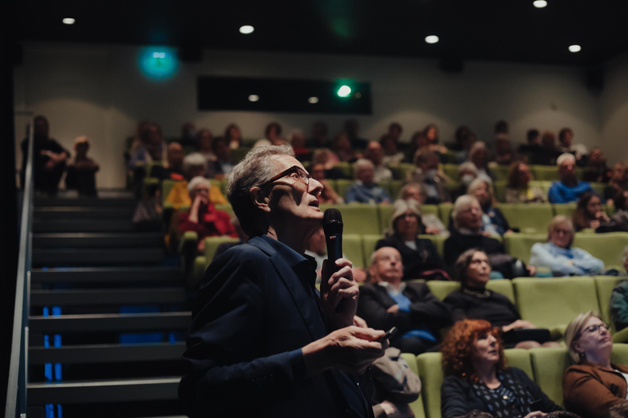Spreker met microfoon houdt een lezing in een goedgevulde zaal met aandachtig luisterend publiek.