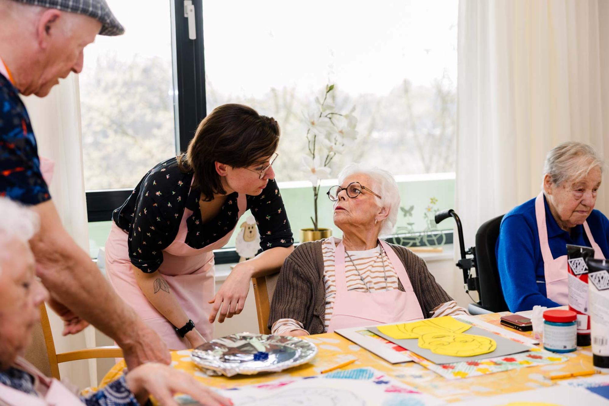 Vier mensen werken samen aan kleurrijke kunstwerken in een lichte ruimte vol creatieve materialen. De sfeer is ontspannen en betrokken.