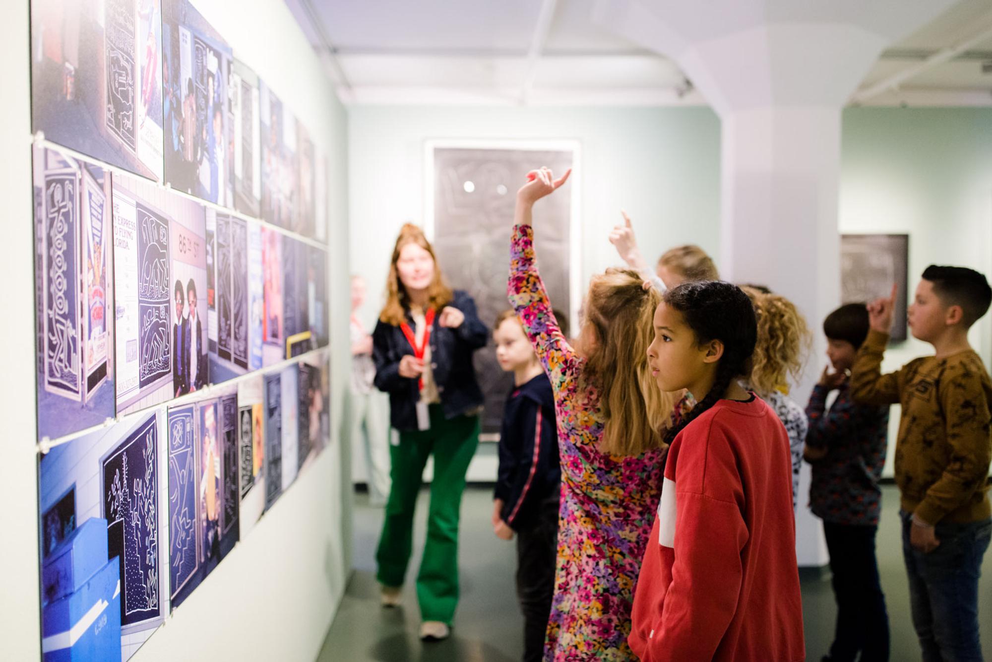 Een groep nieuwsgierige kinderen bekijkt kunstwerken aan de muur tijdens een rondleiding in een museum. Verschillende kinderen steken hun hand op, terwijl een museumgids uitleg geeft. De sfeer is leergierig en betrokken.