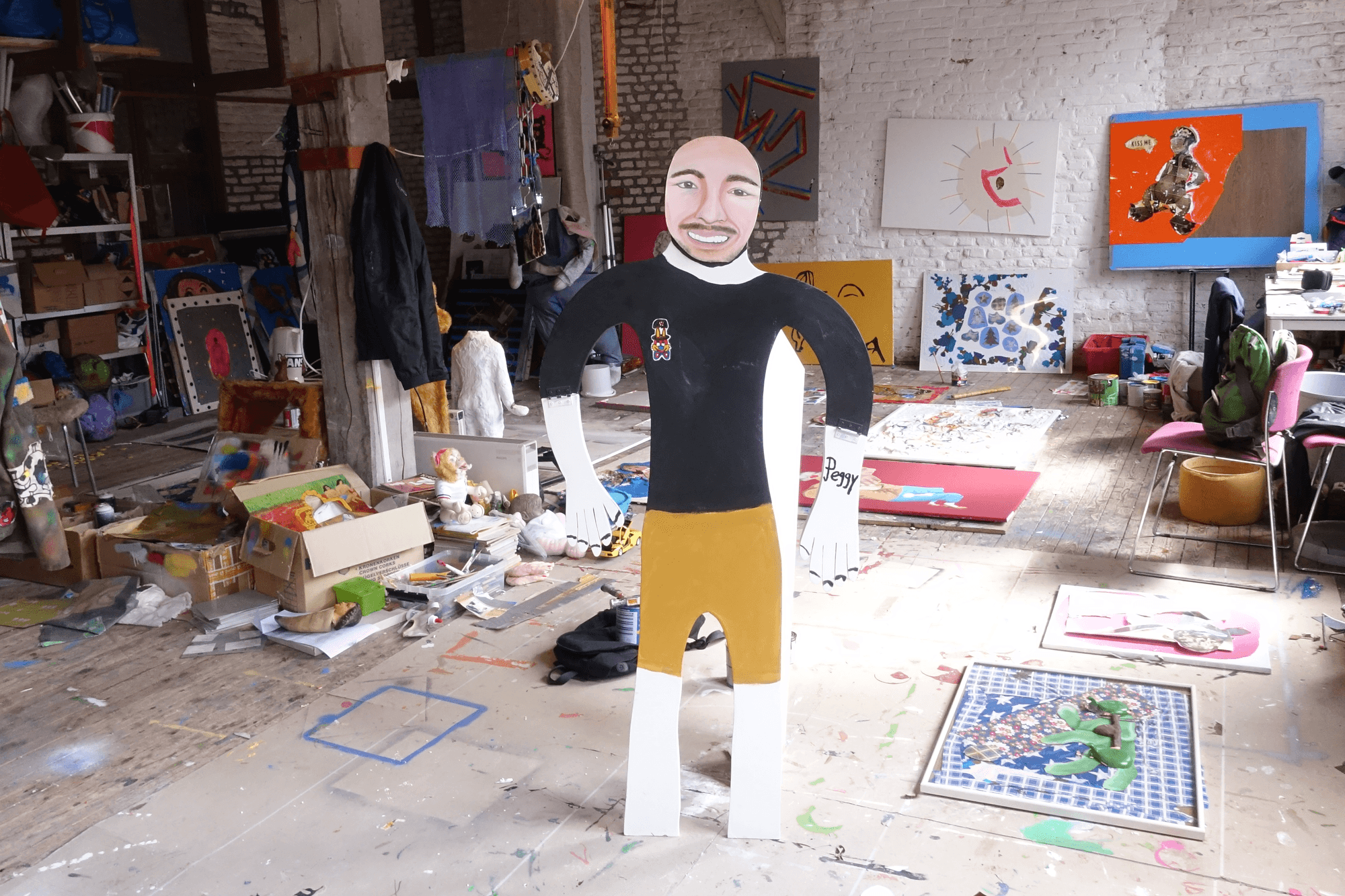 Een kleurrijk en rommelig atelier met schilderijen, kunstmaterialen en dozen op de vloer. In het midden staat een levensgrote, vrolijke kartonnen pop van een man met een zwart shirt en gele broek.