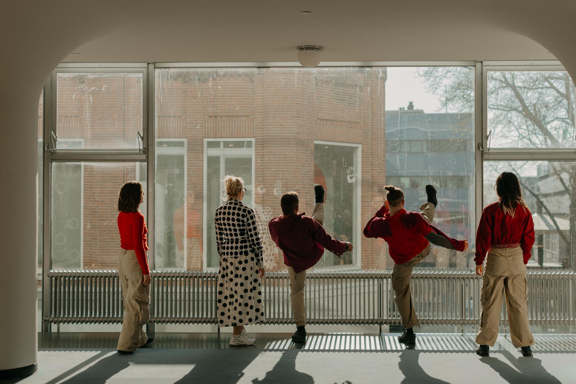 Groep mensen die in een lichte binnenruimte voor een groot raam staan, waarbij enkele personen een choreografische beweging uitvoeren.