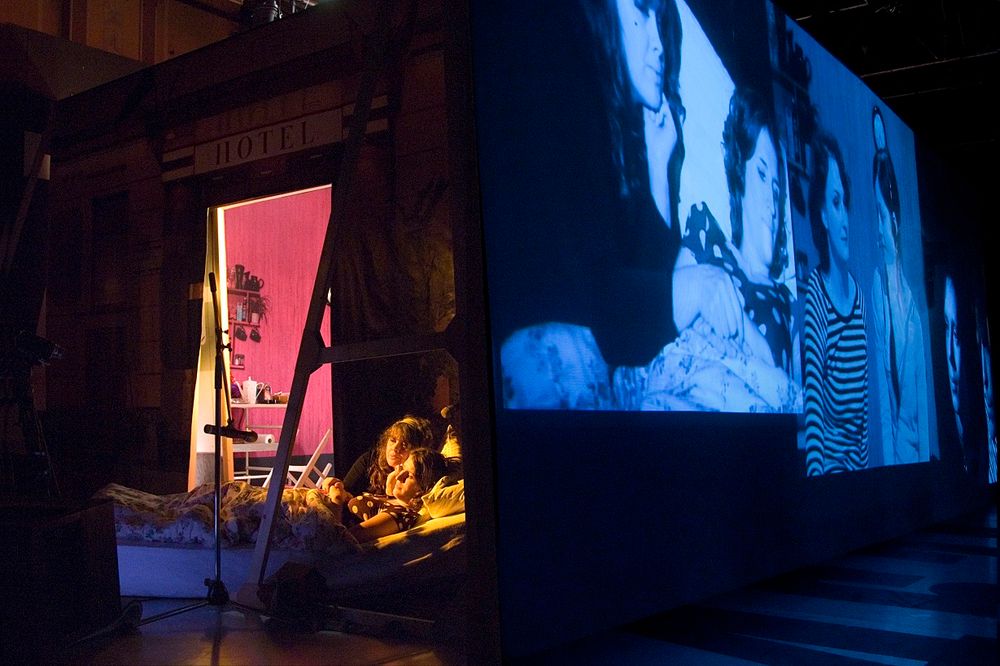 Theaterdecor waar links twee mensen in bed liggen en rechts een videoprojectie te zien is. 