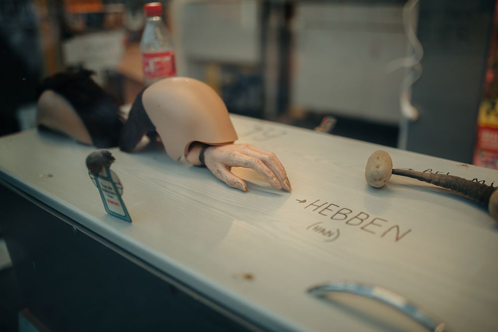 Close-up van een vitrine met onder andere een afgehakte poppenarm, een halve pruik, een flesje cola en het woord ‘HEBBEN’ op een houten plank, onderdeel van een kunstinstallatie in SCHUNCK.