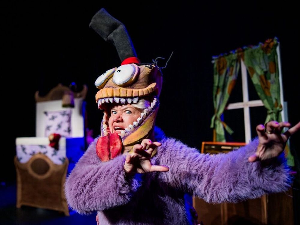 Persoon in een paars harig kostuum met een groot fantasiedierenmasker staat op een theaterpodium tussen decorstukken en een raamdecor.