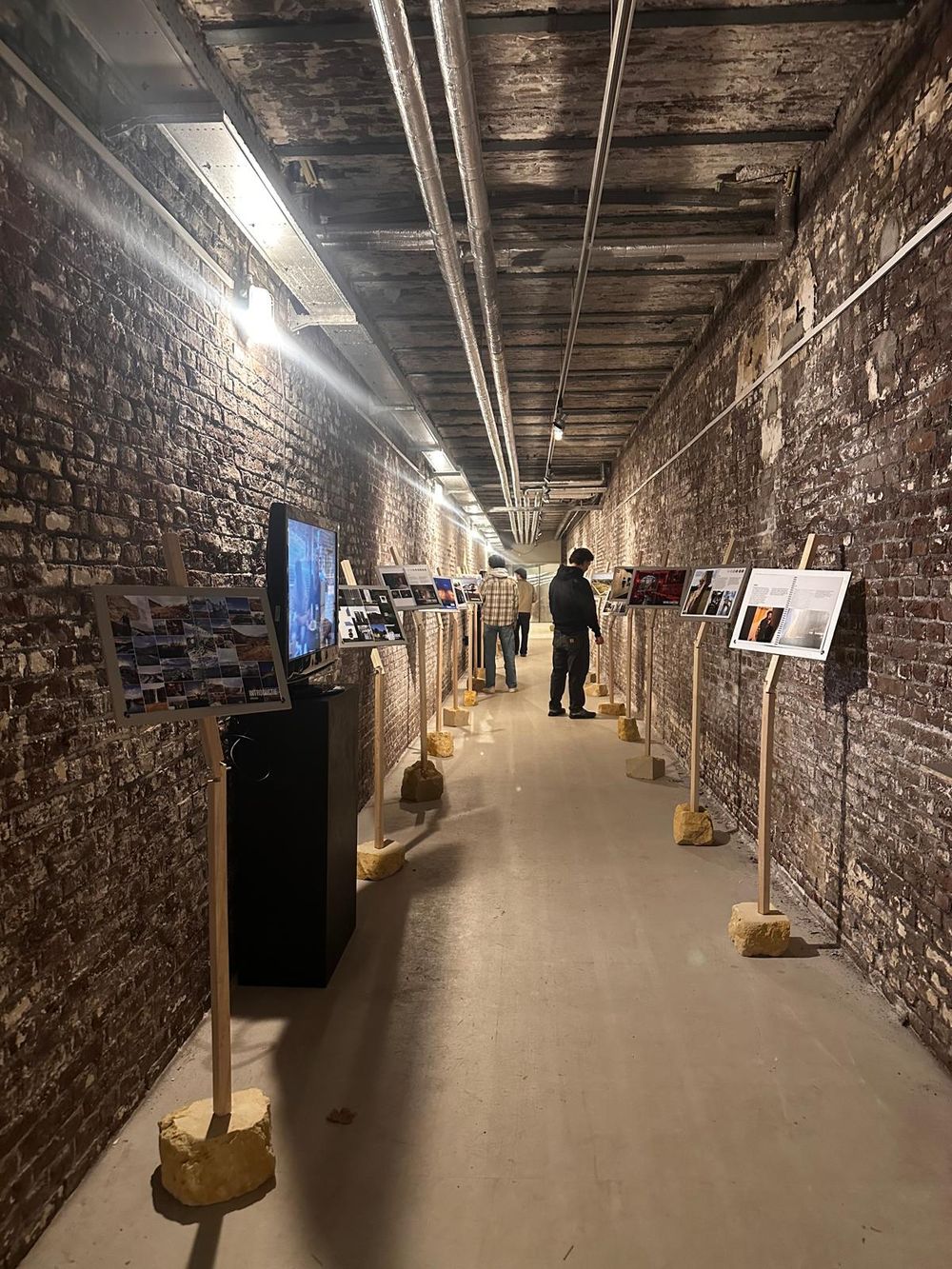 Smalle, lange gang met ruwe bakstenen muren en industriële leidingen, ingericht als fototentoonstelling met beelden op houten standaards; enkele bezoekers lopen en kijken.
