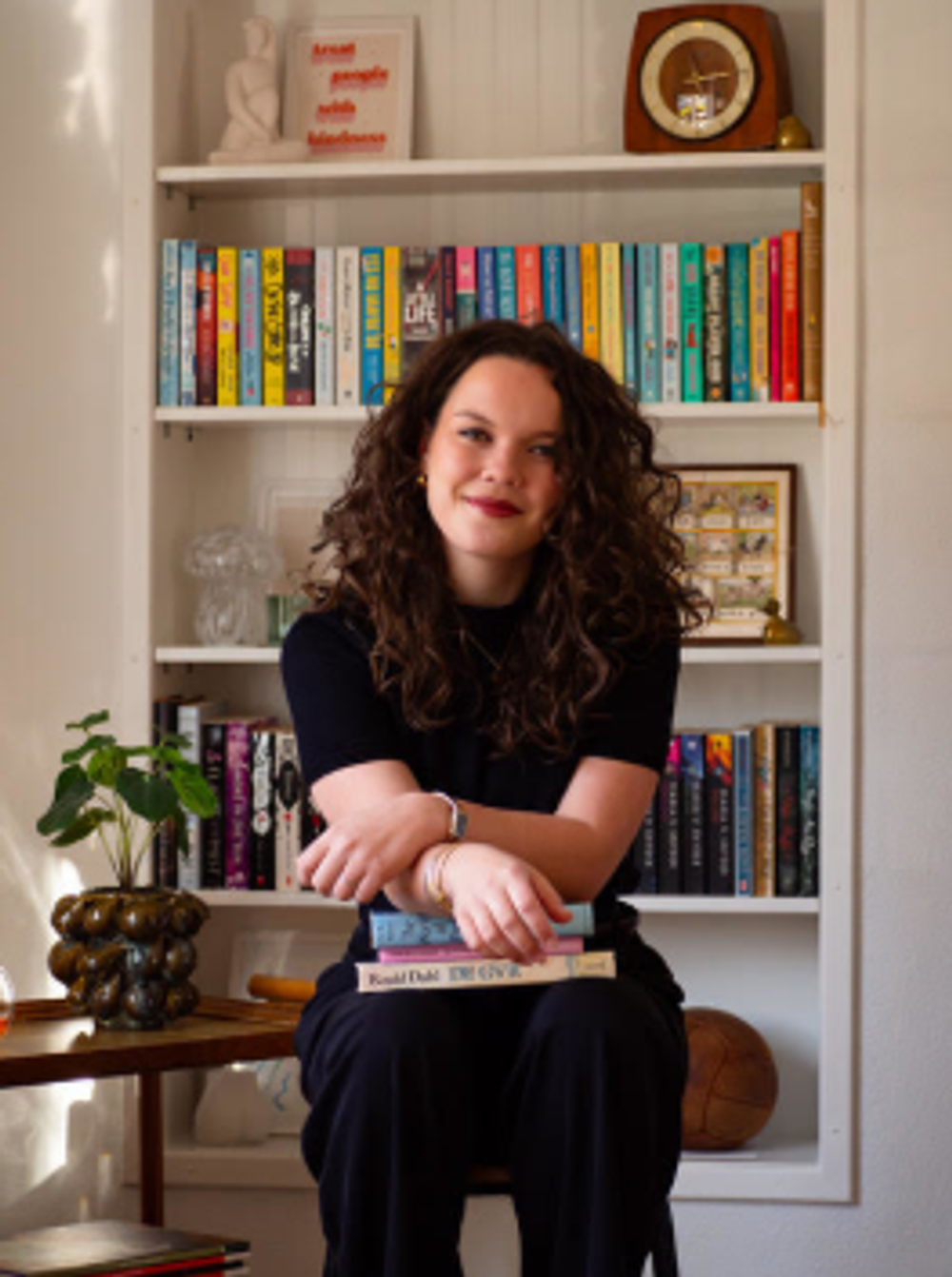 Persoon met krullend haar zit voor een boekenkast gevuld met kleurrijke boeken en houdt een stapel boeken op schoot. De foto is genomen in een huiselijke omgeving met warme verlichting, een plant op een bijzettafel en decoratieve voorwerpen op de planken.
