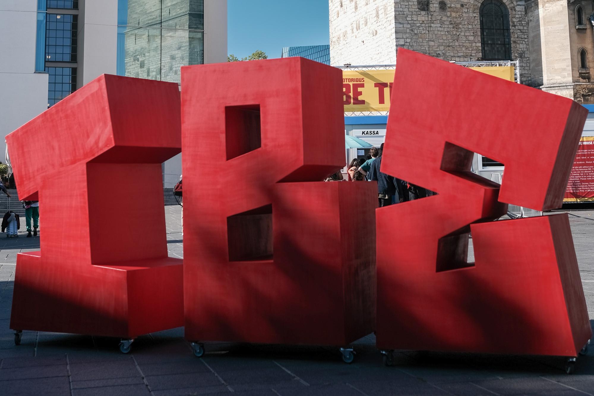 Grote, rode 3D-blokken in de vorm van de letters "I", "B" en "E", geplaatst op wieltjes op een plein voor moderne en historische gebouwen, met mensen op de achtergrond.