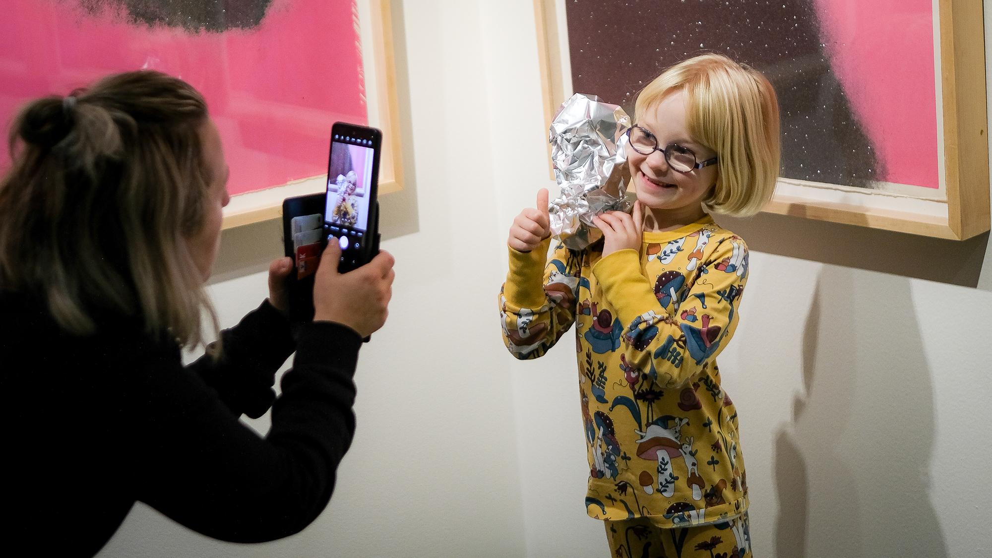 Een kindje dat voor een kunstwerk staat en kijkt naar een jonge vrouw die met haar mobiel een foto van haar maakt, terwijl ze een (zelfgemaakt) kunstwerkje van aluminiumfolie op haar schouder laat leunen en met haar andere hand haar duim omhoog steekt. 