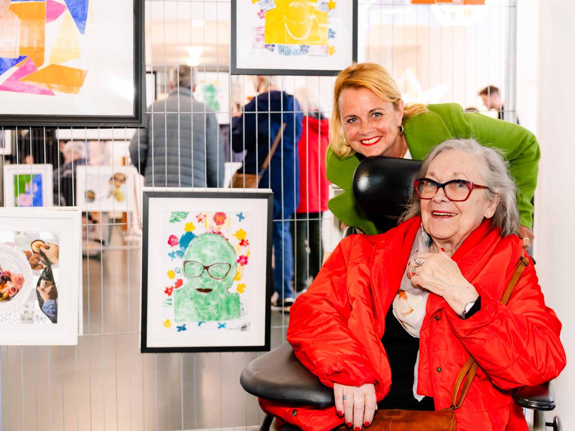 Eindpresentatie van kunstwerken gemaakt door deelnemers aan een kunstproject. Twee mensen staan bij de werken: een persoon in een rolstoel met rode jas en grijs haar, en een begeleider met groene jas. Op de achtergrond hangen kleurrijke kunstwerken aan een hek.