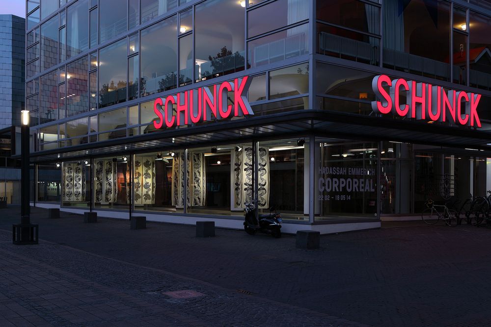 Een modern gebouw met grote glazen ramen en het woord 'SCHUNCK' in rode letters. Er is een overdekte wandelgang en fietsen bij de ingang. Goed verlicht.