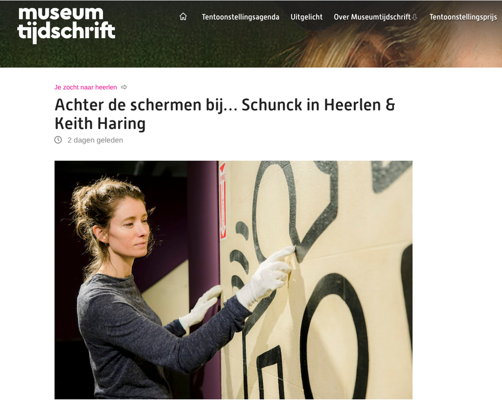 Vrouw met witte handschoenen werkt zorgvuldig aan een kunstwerk met zwarte Keith Haring-lijnen op een muur; bovenaan staat de titel "Achter de schermen bij… Schunck in Heerlen & Keith Haring" op de website van Museumtijdschrift.