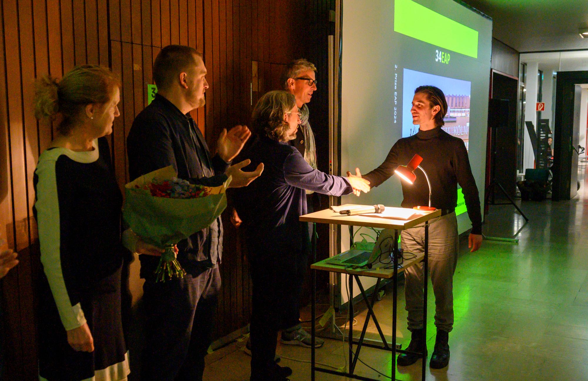Een persoon ontvangt een handdruk en felicitaties tijdens een prijsuitreiking, met op de achtergrond een geprojecteerde presentatie van de Euregional Prize for Architecture. Andere aanwezigen kijken toe en applaudisseren; iemand houdt een boeket bloemen vast.