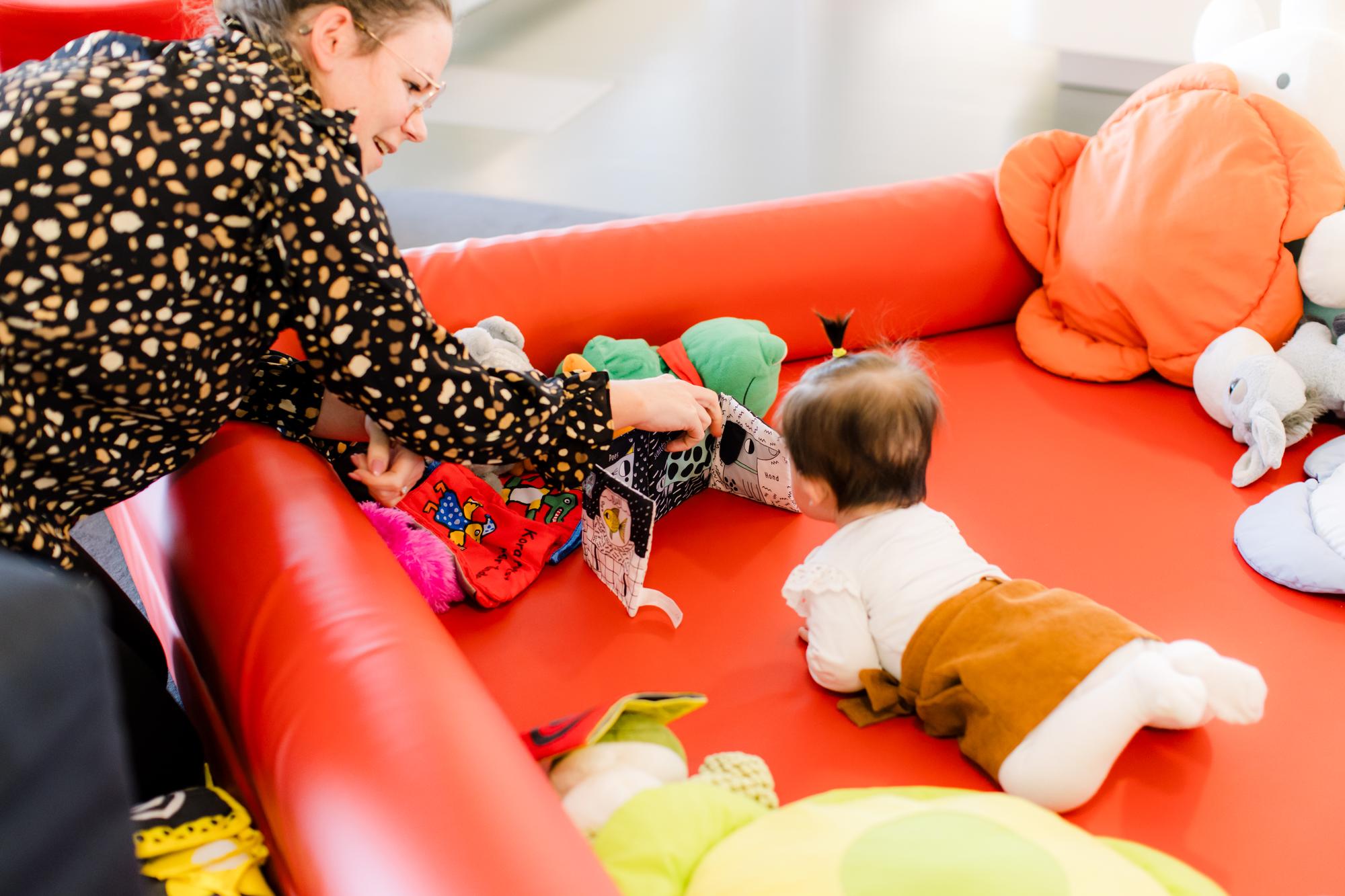 Een baby ligt op de buik in een rood speelkleed terwijl een volwassene samen met het kind een stoffen prentenboek bekijkt, omringd door kleurrijke knuffels en speelgoed.