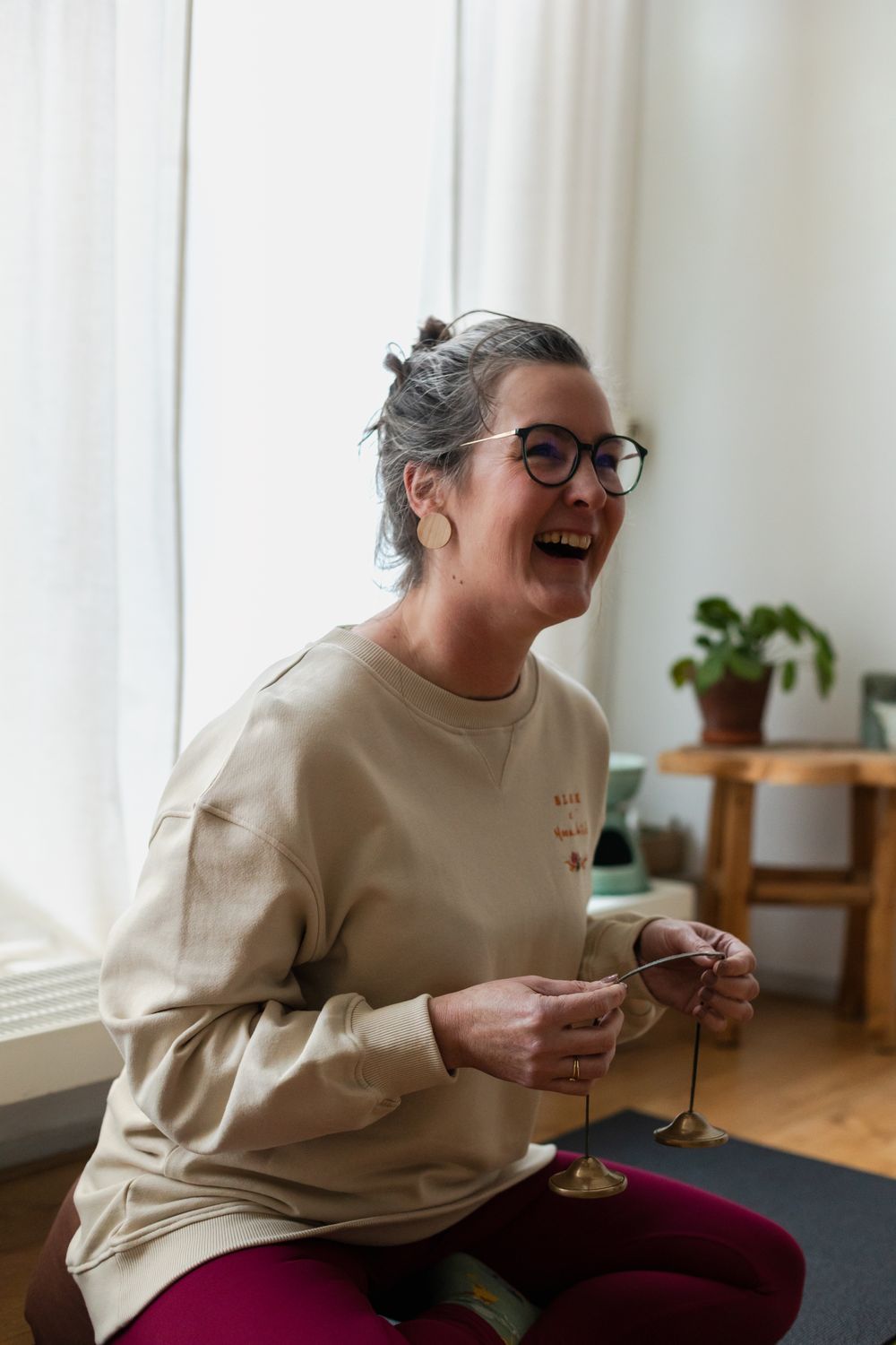 Lachende vrouw met bril zit in kleermakerszit op een yogamat binnen, en houdt een set tingsha-belletjes vast. Ze draagt een beige trui en paarse leggings; op de achtergrond staan een houten tafeltje en een plant.