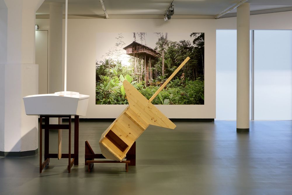 Houten object in het museum met op de achtergrond een foto van een hoge hut in een jungleachtige omgeving, hangend aan de wand in het museum.