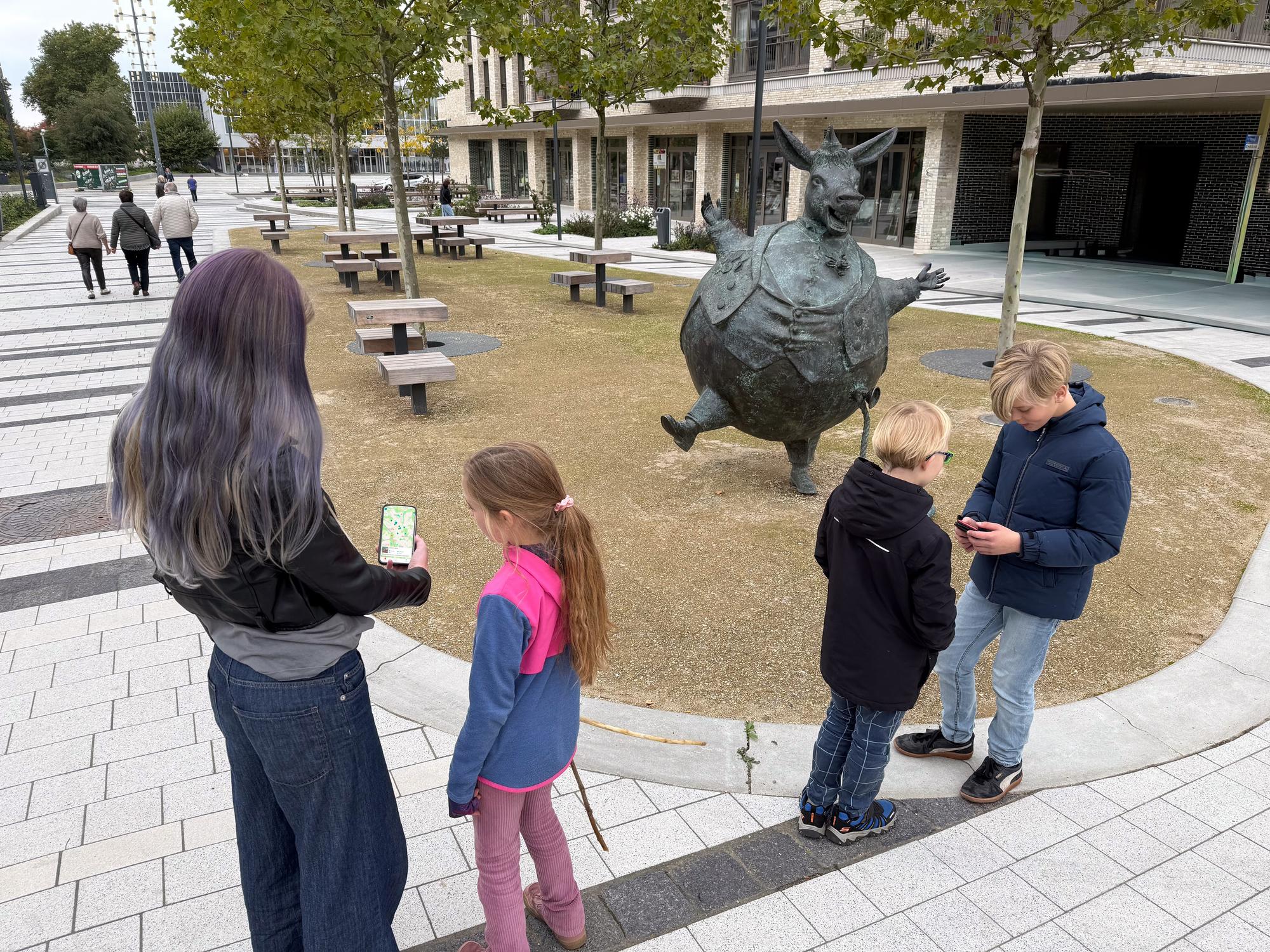 Een groep kinderen staat op een plein bij een speels bronzen beeld van een dier op twee poten. Twee kinderen kijken op een telefoon, terwijl op de achtergrond mensen over de promenade lopen tussen bomen en bankjes.