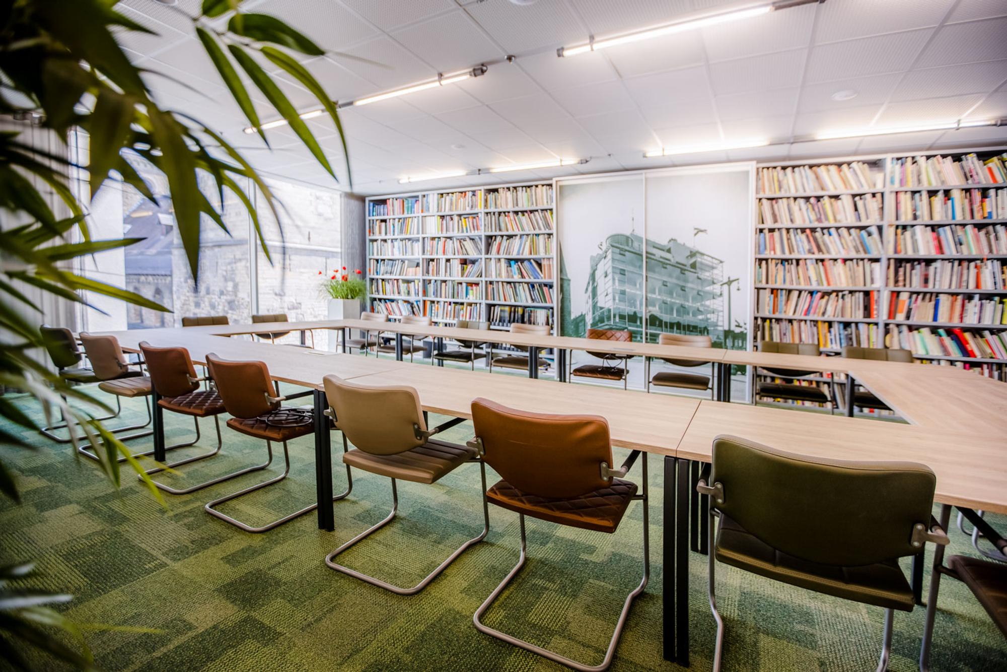  Vergaderzaal tafels in een carré met stoelen, wand met boeken op de achtergrond. 