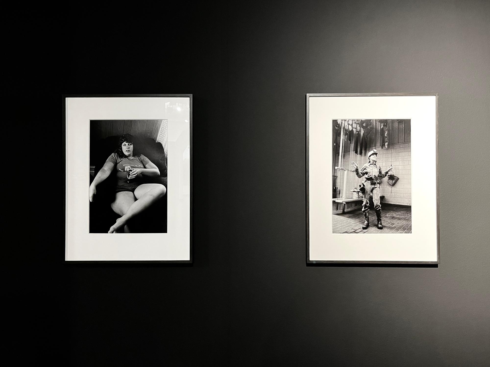 Twee ingelijste zwart-witfoto's aan een donkere muur in een tentoonstellingsruimte; links een zittende vrouw met een blikje in de hand, rechts een man in uniform met werkgerei om zich heen.