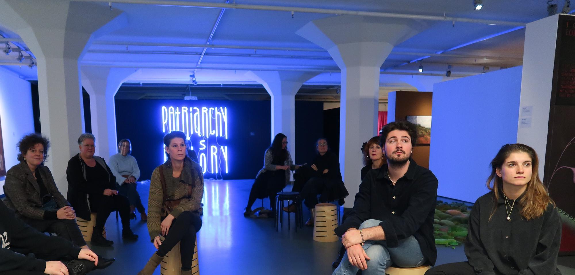 Leden van de Badeklasse zitten op krukken in de museumzaal, voor hen staat kunstenaar David Bade. Op de achtergrond is het kunstwerk 'Patriarchy is History' te zien, gemaakt van blauwe neonbuizen.